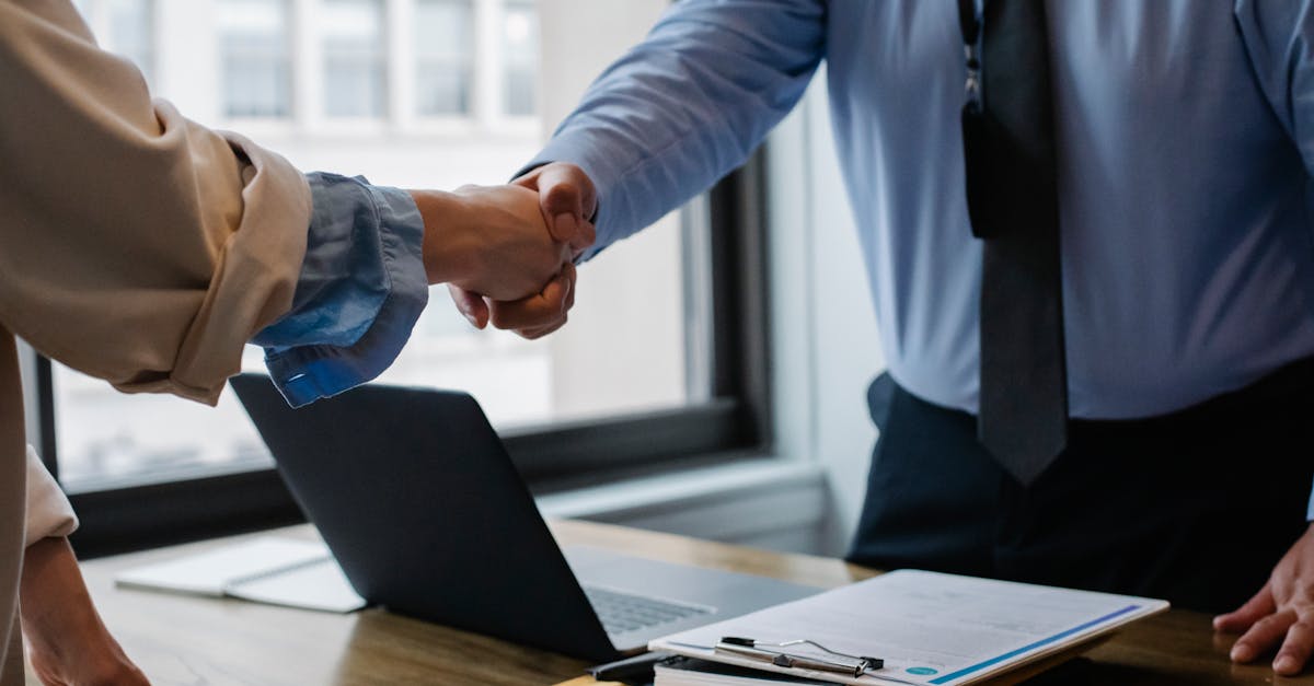 Crop unrecognizable coworkers in formal wear standing at table with laptop and documents while greeting each other before meeting